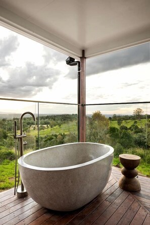 Eine große, freistehende Badewanne auf einer Holzterrasse mit Glasgeländern mit Blick auf eine Landschaft.