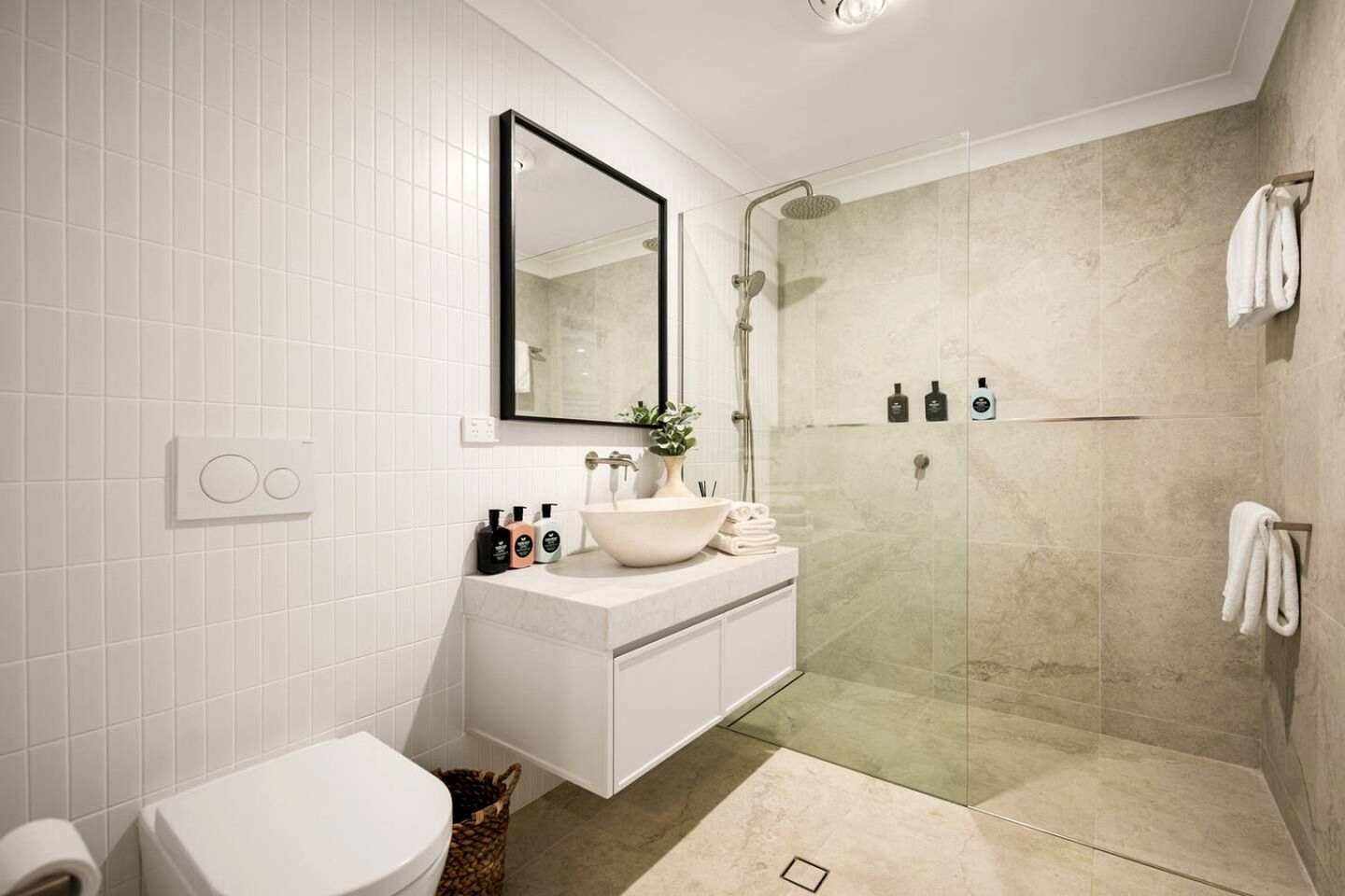 A modern bathroom with a white tiled wall, a large mirror, a white sink, a glass shower enclosure, and a toilet.