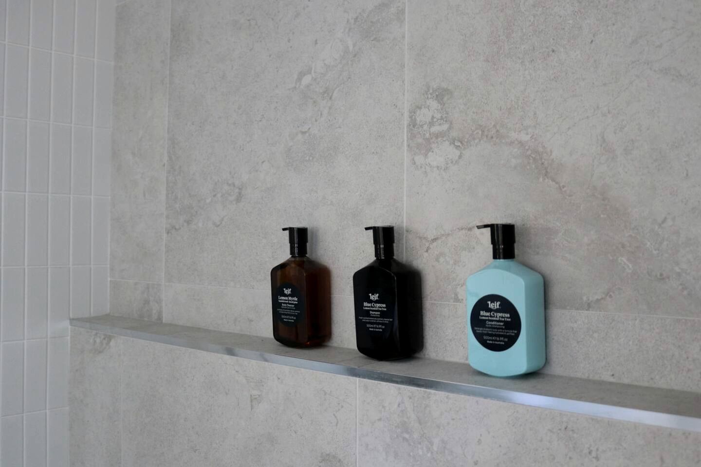 Three bottles of body care products on a shelf against a tiled wall.