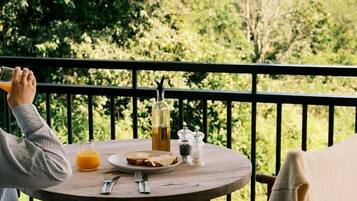 A person sitting at a round wooden table with a glass of orange juice, a bottle, and a plate of food on a balcony with a view of trees and hills.