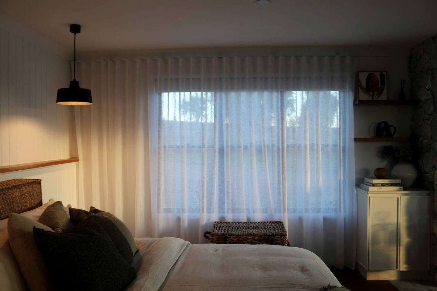 Un dormitorio con cama, mesita de noche, lámpara, ventana con cortinas translúcidas y un pequeño banco.