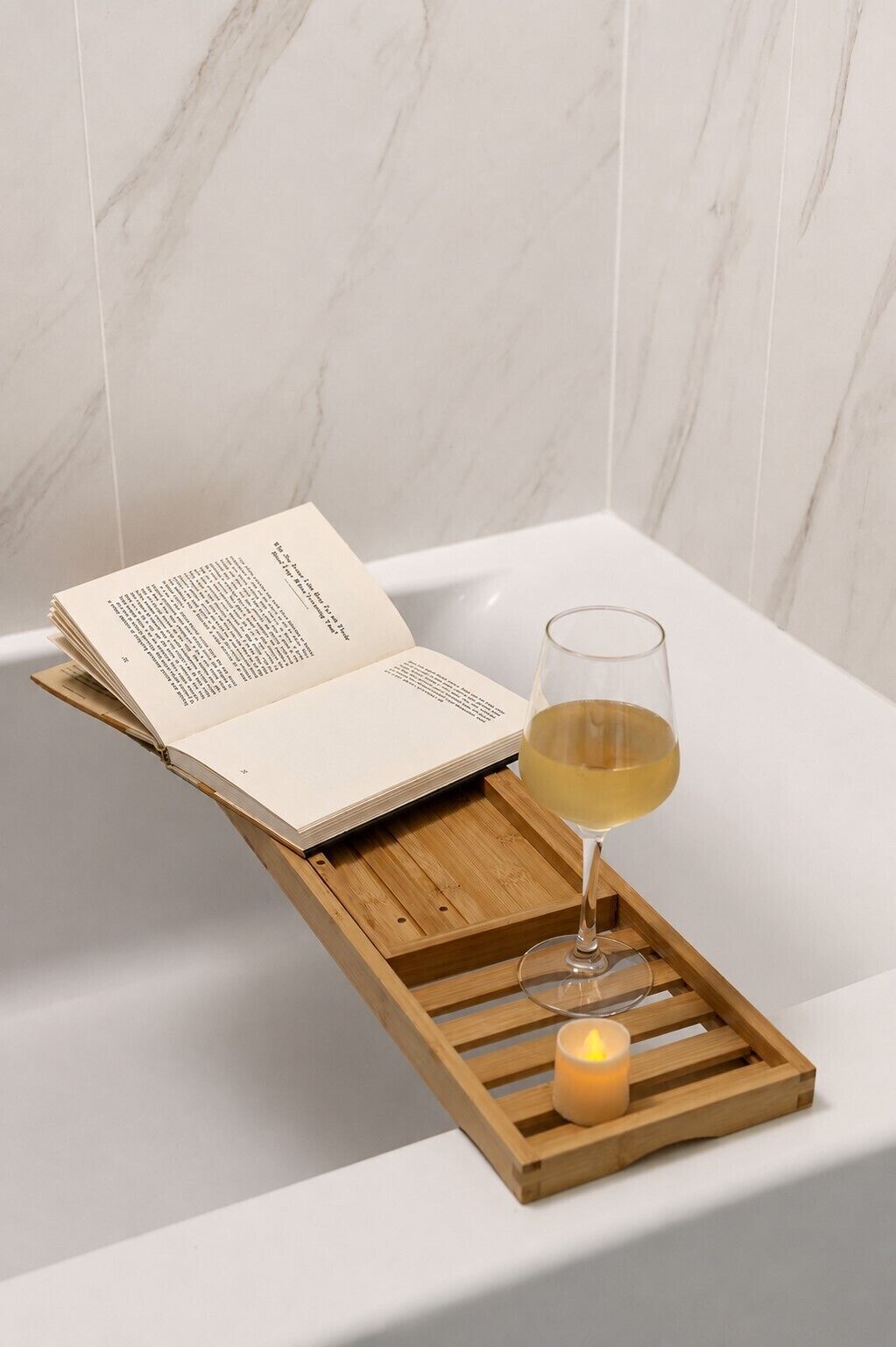 A book, a glass of white wine, and a lit candle on a wooden tray in a bathtub.