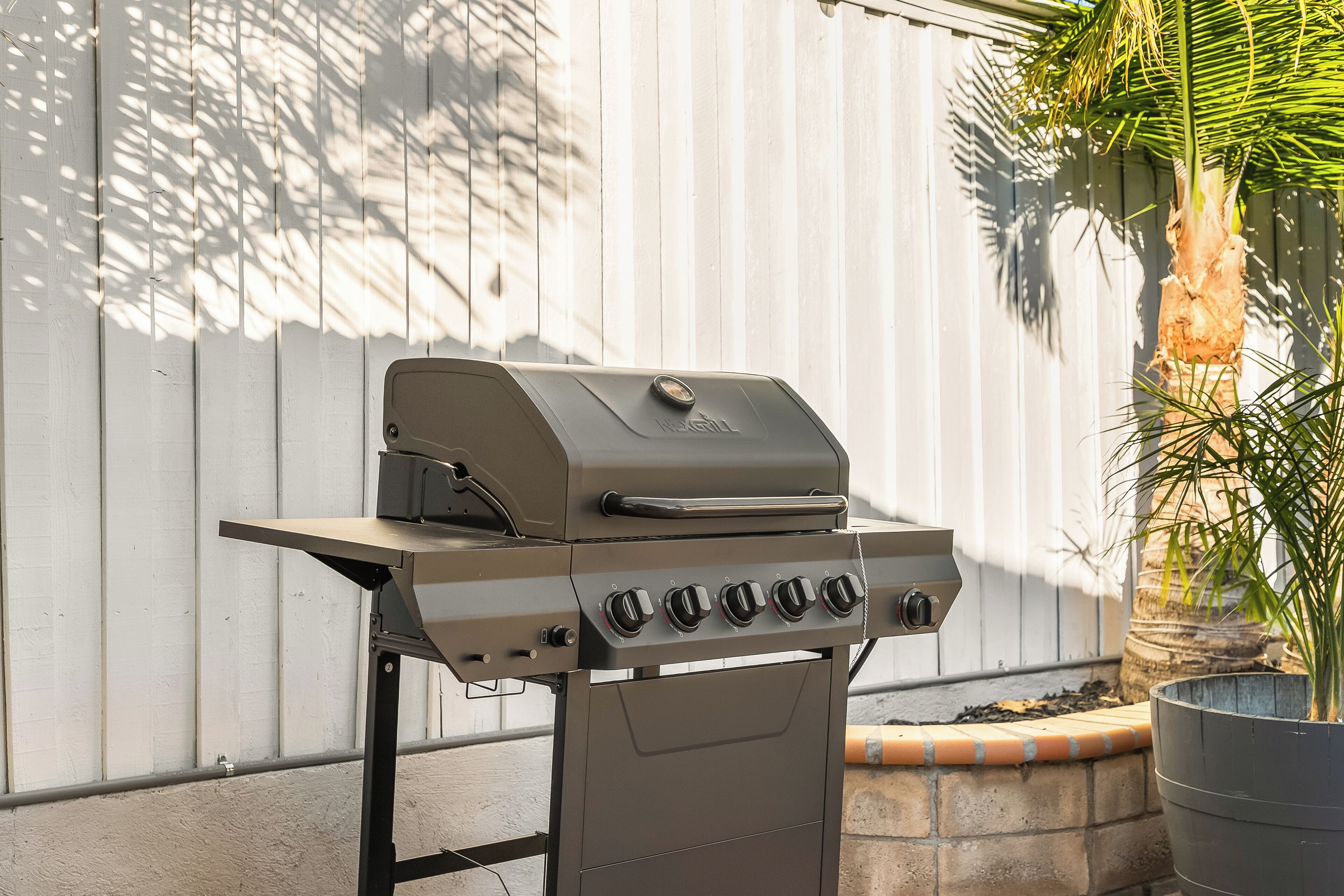 A barbecue grill with a control panel and a lid, positioned against a white wall with shadows cast by nearby foliage.