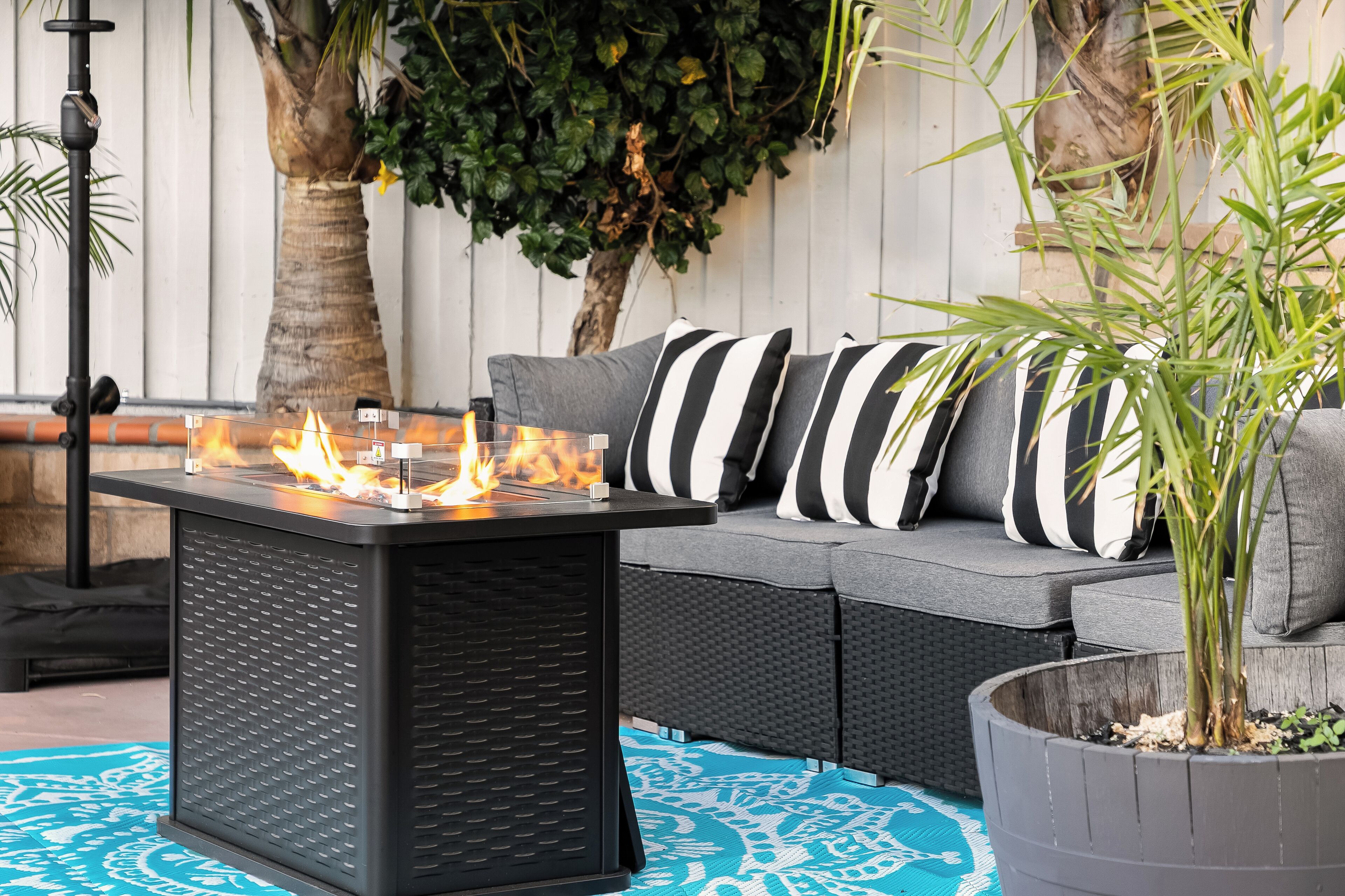 A patio with a black fire table, grey sofa with striped pillows, and a potted plant.