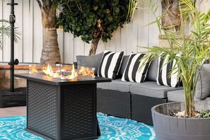 A patio area with a black metal table with a glass top, a gray sofa with striped pillows, and a potted plant.