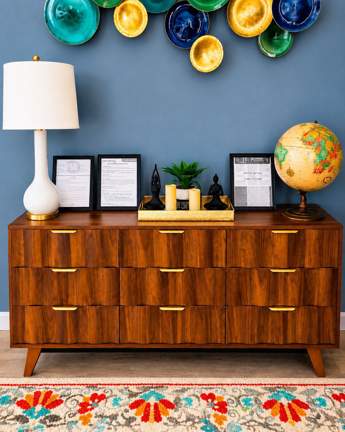 A wooden dresser with multiple drawers, a white lamp, framed certificates, a small plant, candles, and a globe.