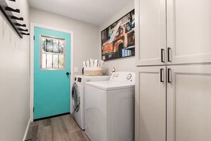 Een wasserette met een turquoise deur, een wasmachine, een droger en een schilderij van een oldtimer vrachtwagen aan de muur.
