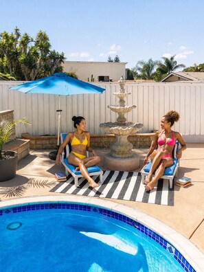 Duas pessoas sentadas à beira da piscina, perto de uma fonte, em um espaço cercado por um muro branco e palmeiras.