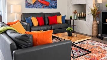 A modern living room with a black leather sofa, colorful cushions, a large painting of boats, a marble wall, and a patterned rug.