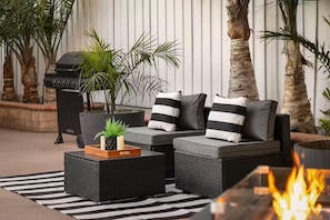 A sectional sofa with striped pillows, a small potted plant, a grill, and a fire feature.