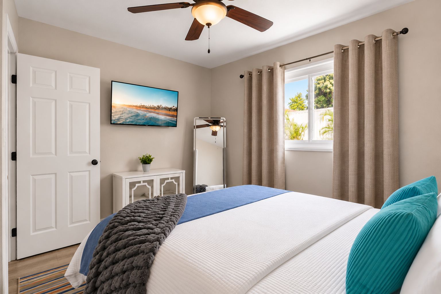 A bedroom with a bed, a ceiling fan, a window with curtains, a bedside table with a lamp, and a wall-mounted TV.