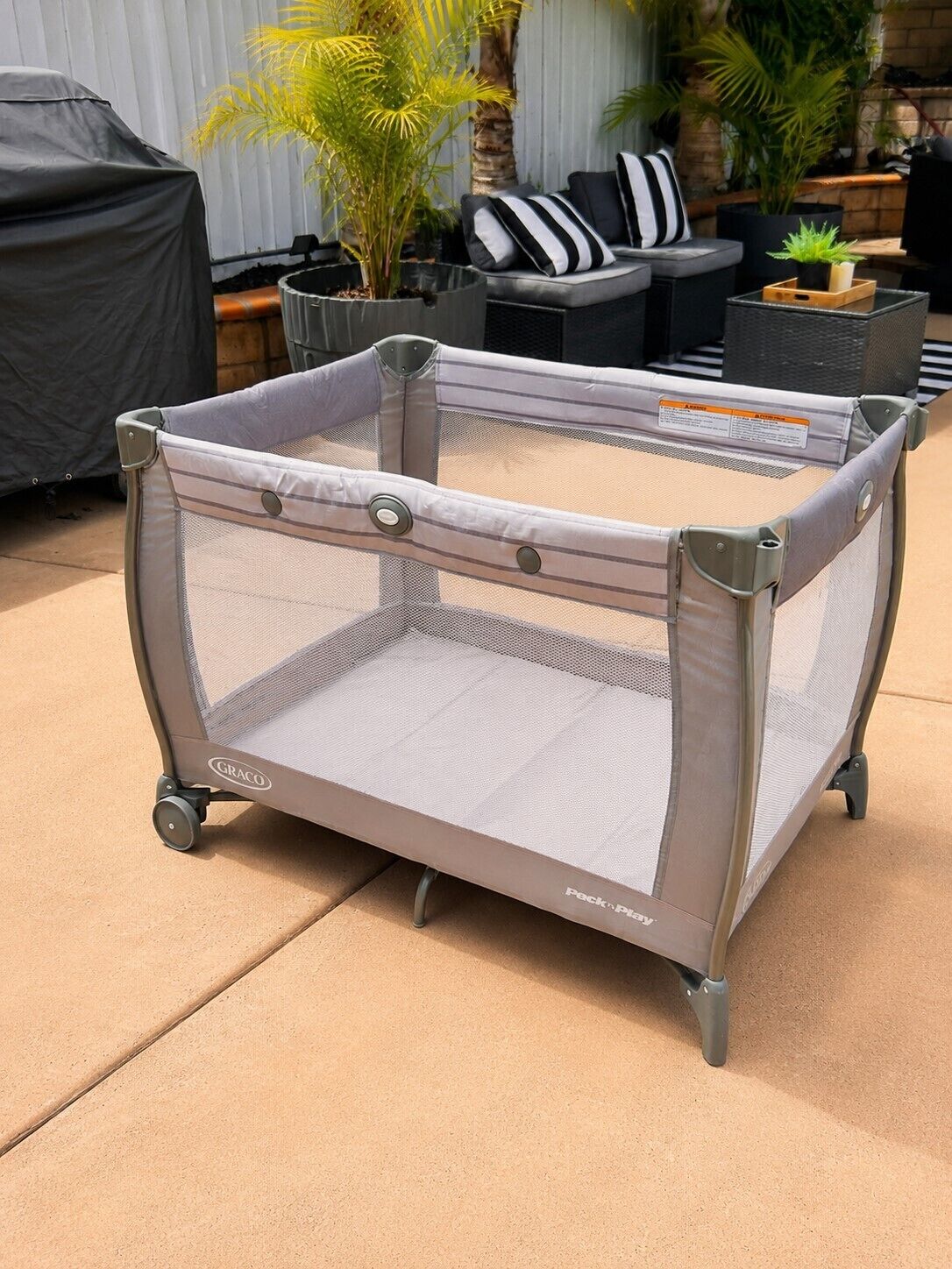A portable baby playpen with wheels, labeled 'GRACO' and 'Peck & Play,' on a concrete patio.