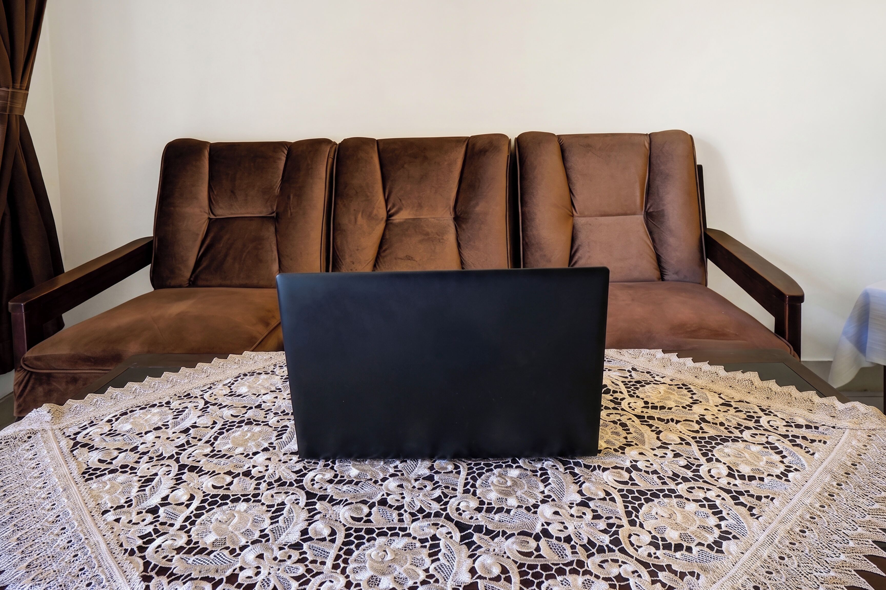 Una laptop sobre una mesa con mantel de encaje, un sofá marrón y una cortina en una habitación.
