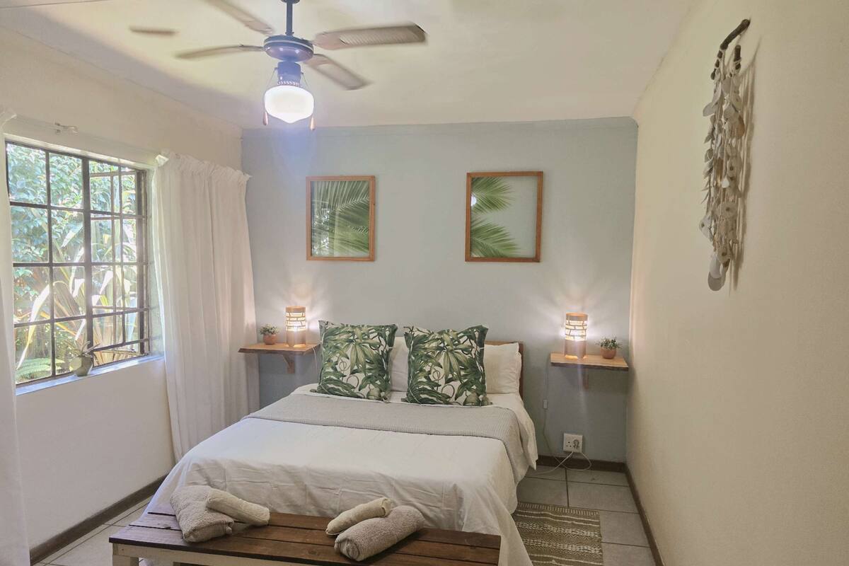 A bedroom with a bed, a ceiling fan, two wall-mounted lamps, a window with curtains, and a small table with towels.