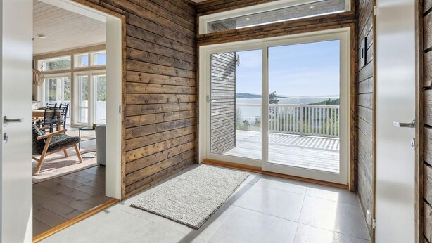 Una habitación con pared de madera, puerta corrediza de vidrio que da a un balcón con vista a una masa de agua, y un comedor con sillas y una mesa.