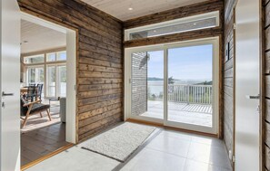 Una habitación con pared de madera, puerta corrediza de vidrio que da a un balcón con vista a una masa de agua, y un comedor con sillas y una mesa.