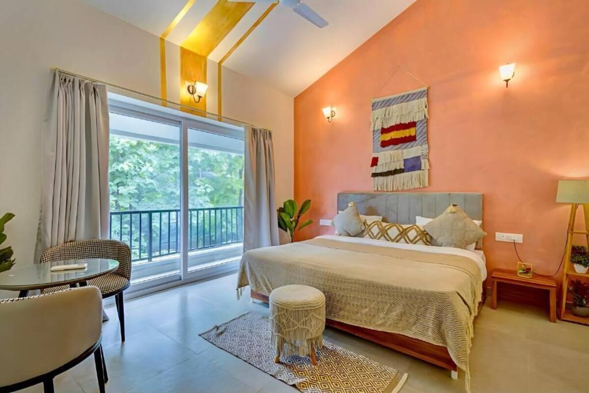 Un dormitorio con cama, una mesita, una silla, una ventana con cortinas y una lámpara fijada a la pared.