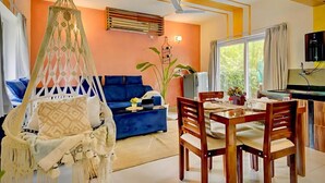 Una sala de estar con un sofá azul, una mesa de comedor de madera con sillas, una silla colgante y un ventanal grande.