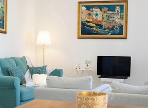 A living room with a teal sofa, a white lamp, a TV, and a painting of a coastal scene. - Infinite Blue View-Alghero (Alghero)