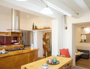 Eine moderne Küche mit Dunstabzugshaube aus Edelstahl, einem eingebauten Backofen und einer Holz Arbeitsplatte. Es gibt einen Essbereich mit Tisch und Stühlen sowie ein Wohnzimmer mit Sofa und Spiegel.