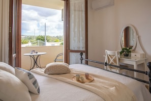 Ein Schlafzimmer mit Bett, Nachttisch, Sessel und kleinem Tisch mit Tassen, mit Blick ins Freie.