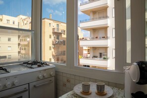 Eine Küche mit einem Gasherd, einem Fenster mit Blick auf Wohnhäuser und einem Tisch mit zwei Tassen.
