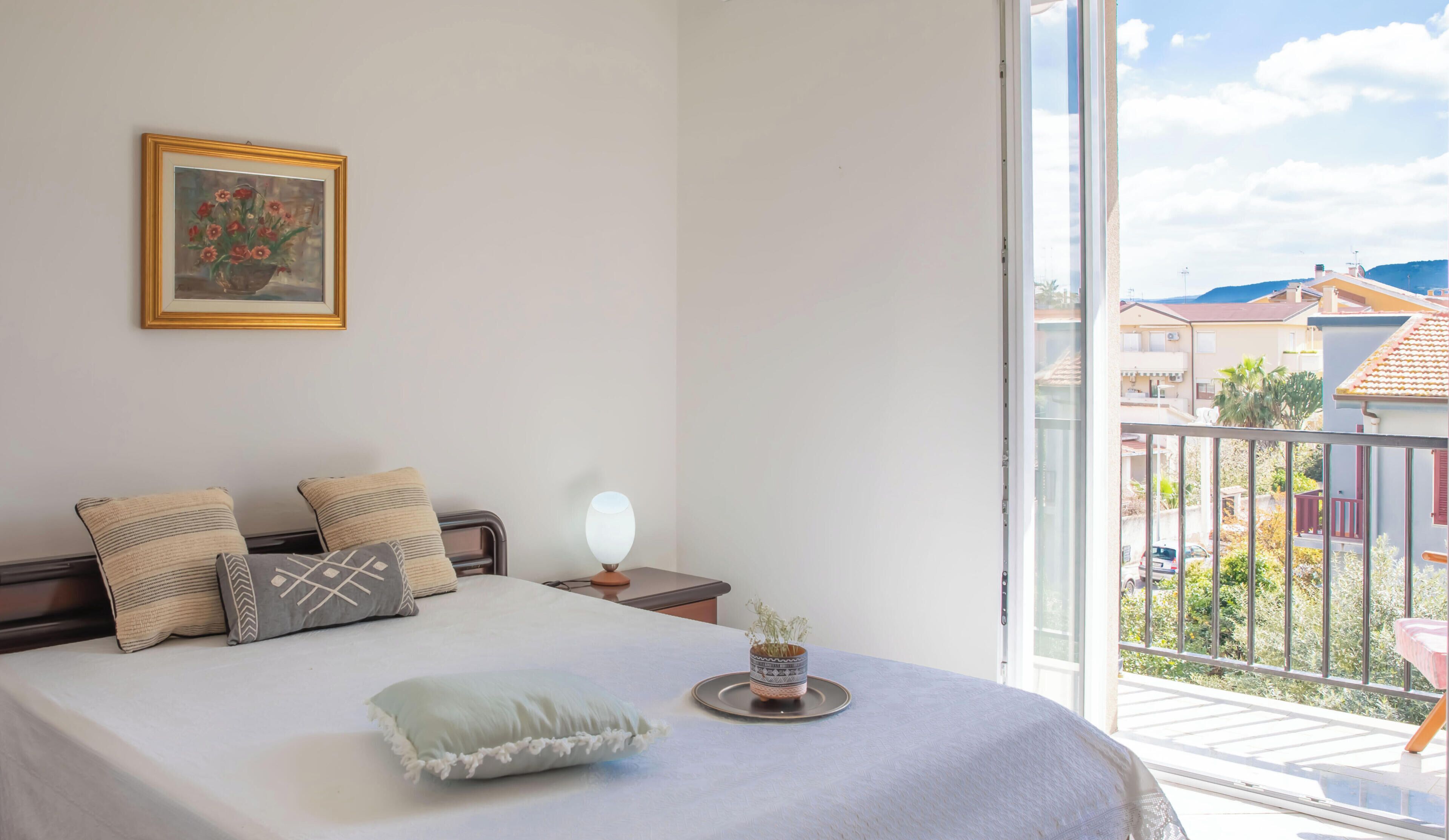 A bedroom with a bed, pillows, a bedside table with a lamp, a small potted plant, and a window with a view of buildings.