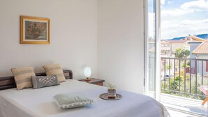 Une chambre à coucher avec un lit, des oreillers, une table de chevet avec une lampe, une petite plante en pot et une fenêtre donnant sur des immeubles.
