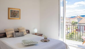 Ein Schlafzimmer mit Bett, Kissen, einem Nachttisch mit Lampe, einer kleinen Topfpflanze und einem Fenster mit Blick auf Gebäude.