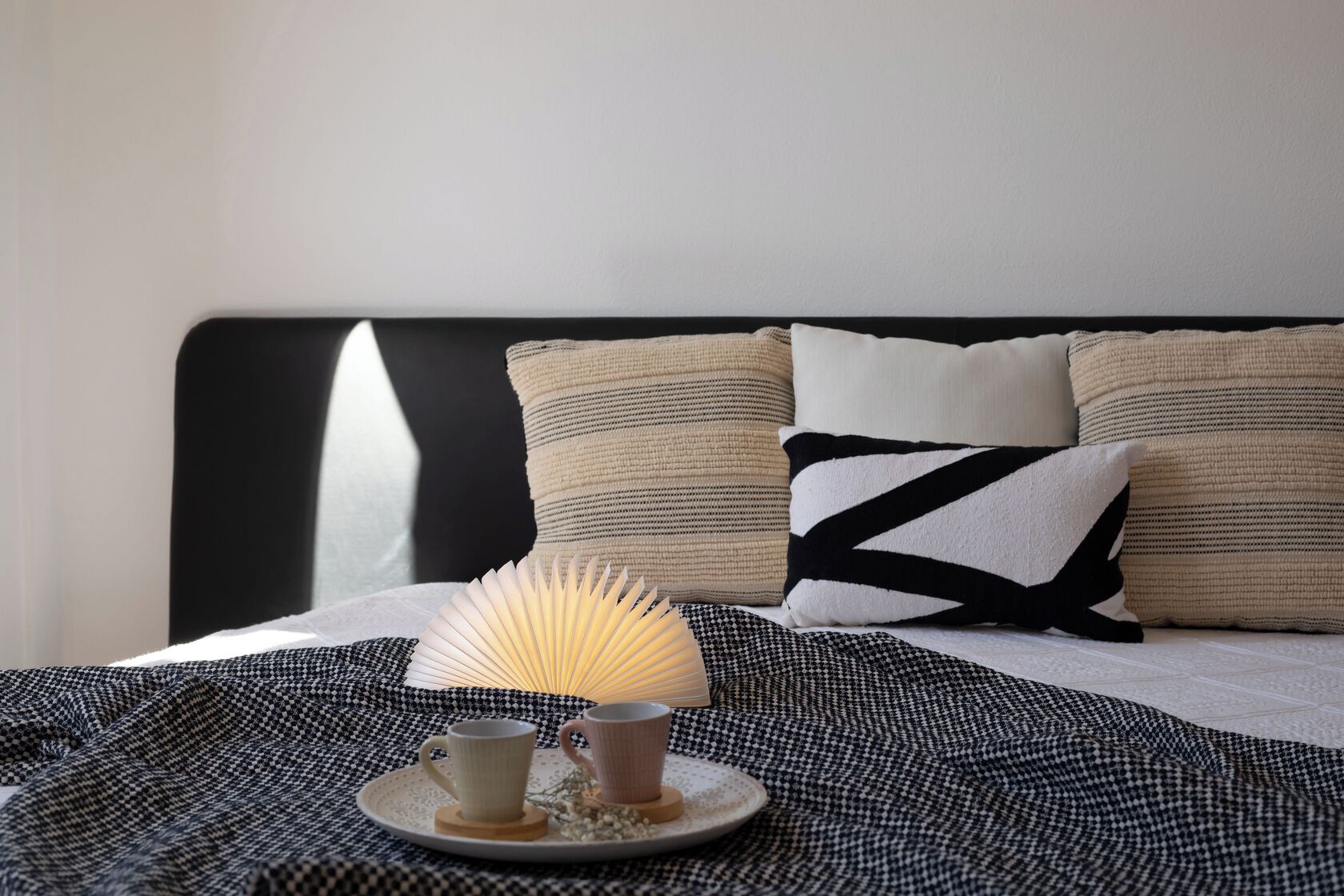 Een net opgemaakt bed met een zwart hoofdeinde, een bedlamp, twee bekertjes en een bord met een bloem.