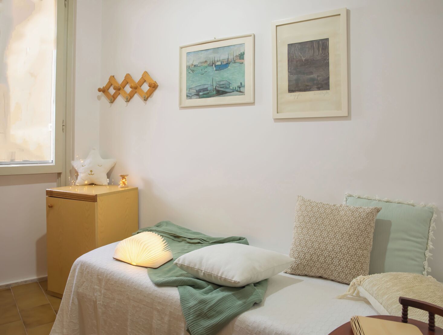 Un dormitorio con cama, almohadas, una mesita de noche, una ventana y dos cuadros en la pared.