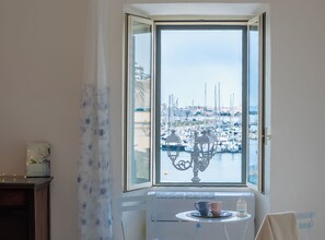 A room with a large window overlooking a marina, a small table with cups, and a radiator. - Suite Porto antico - Alghero (Alghero)