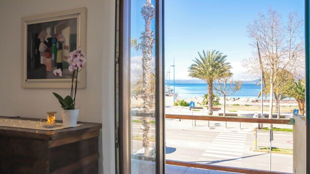 Een kamer met een groot raam dat uitkijkt over een strand, een bureau met een plant en een lijst aan de muur.