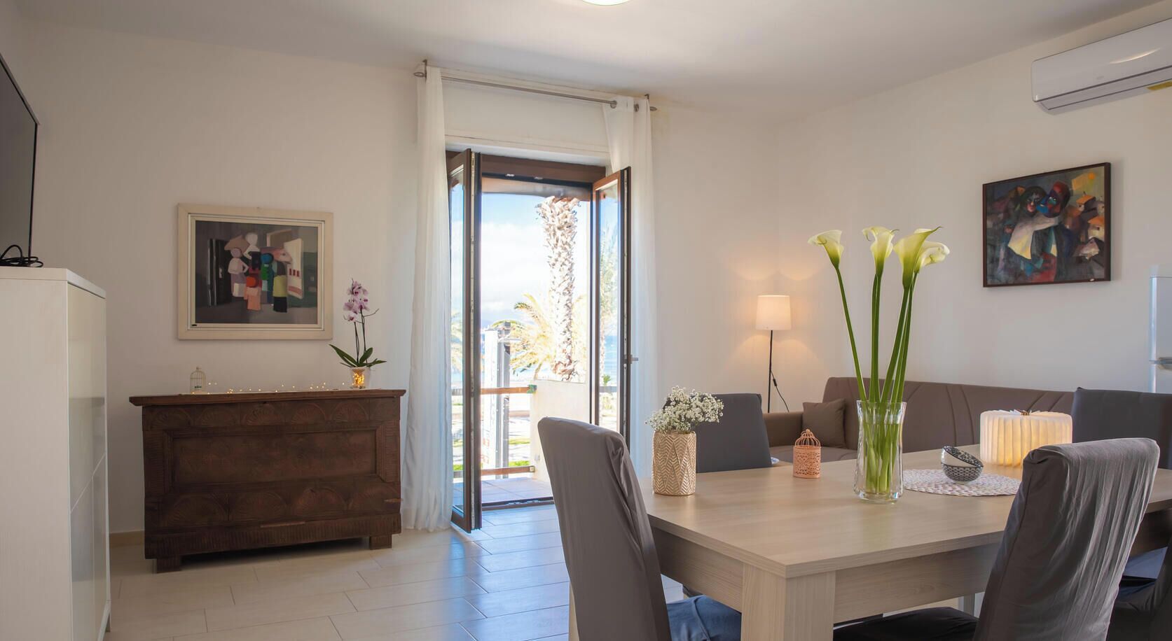 A dining area with a wooden table, chairs, a vase with flowers, a lamp, and a painting on the wall.