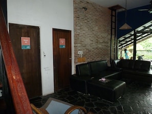 A lobby with two doors labeled 'Room #4' and 'Room #5,' a black leather sofa, a wicker chair, and a stone floor. - Nok Guest House And Hostel (Koh Tao)