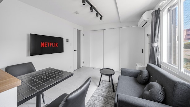 Una sala de estar moderna con un televisor de pantalla plana mostrando 'NETFLIX', una mesa de comedor, un sofá y una ventana con cortinas.