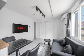 A modern living room with a flat-screen TV displaying 'NETFLIX,' a dining table, a sofa, and a window with curtains. - ASTAGE CHITOSE SUEHIRO 101  (Chitose)