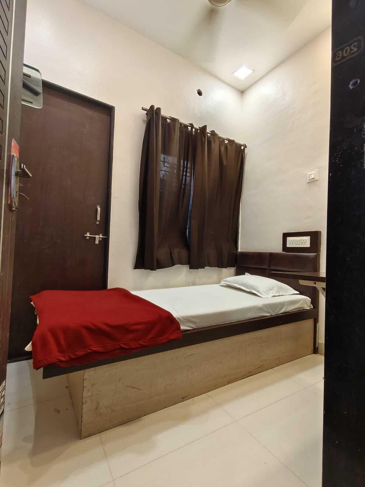 Un lit simple avec une couverture rouge, une tête de lit en bois, un rideau marron, une porte et une petite table de chevet dotée d’une prise électrique.