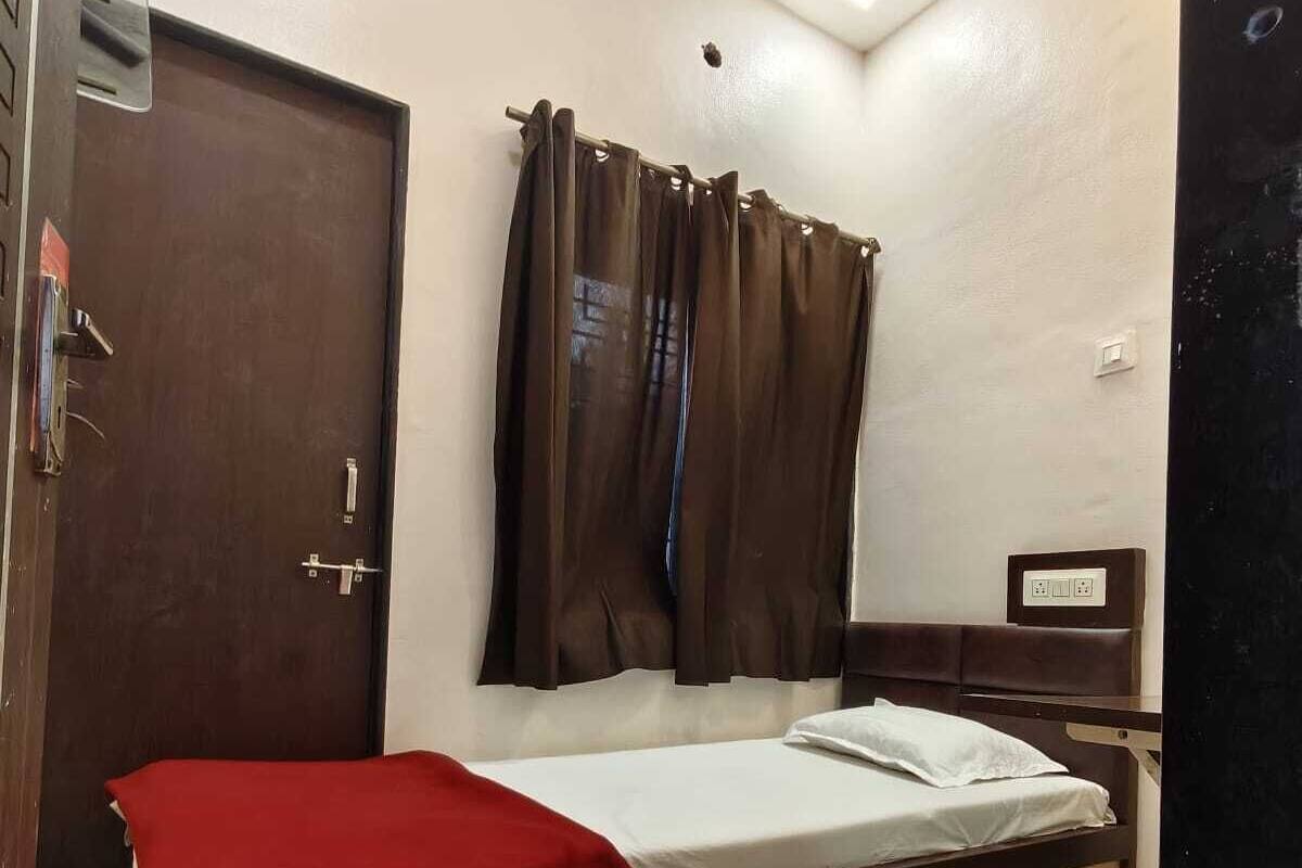 Una cama individual con una manta roja, cabecera de madera, cortina marrón, una puerta y una mesita de noche pequeña con toma de corriente.