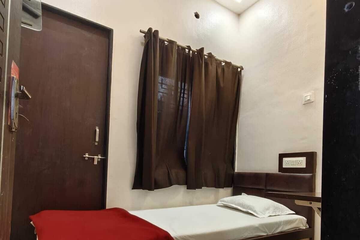 Un lit simple avec une couverture rouge, une tĂȘte de lit en bois, un rideau marron, une porte et une petite table de chevet dotĂ©e dâune prise Ă©lectrique.
