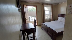 A bedroom with a bed, a desk, and a chair. There is a window with curtains, a door, and a balcony visible. - Crown Accomodation (Narok)