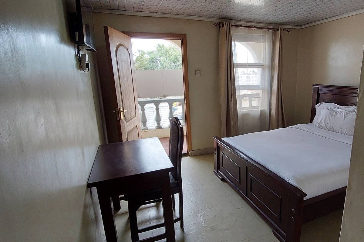 Une chambre à coucher avec un lit, un bureau et une chaise. On y voit une fenêtre avec des rideaux, une porte et un balcon.