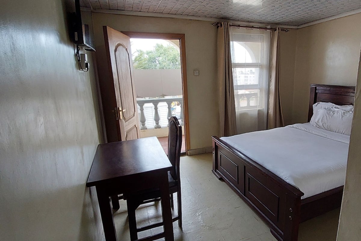 A bedroom with a bed, a desk, and a chair. There is a window with curtains, a door, and a balcony visible.