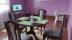A dining area with a round table, chairs, a television displaying a news program, and a window with curtains. - Crown Accomodation (Narok)
