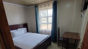 A bedroom with a wooden bed, a small table, and a window with curtains. - Crown Accomodation (Narok)