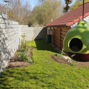 Ein von einer Mauer umschlossener Gartenbereich mit einer Holzkonstruktion, einem grĂŒnen Vogelhaus und einer SteinsĂ€ule.