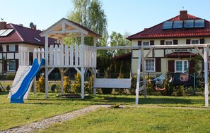 Ein Spielplatz mit Rutsche und Schaukeln, umgeben von einem weißen Zaun, mit Gebäuden im Hintergrund.