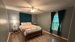 Un dormitorio con una cama de madera, un ventilador de techo, una mesita de noche con lámpara y una ventana con cortinas.