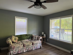 A room with a floral-patterned sofa, a ceiling fan, and two windows with blinds.