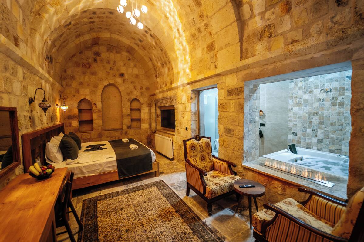A stone-walled room with a bed, a chair, a small table, and a bathtub with a fire feature.